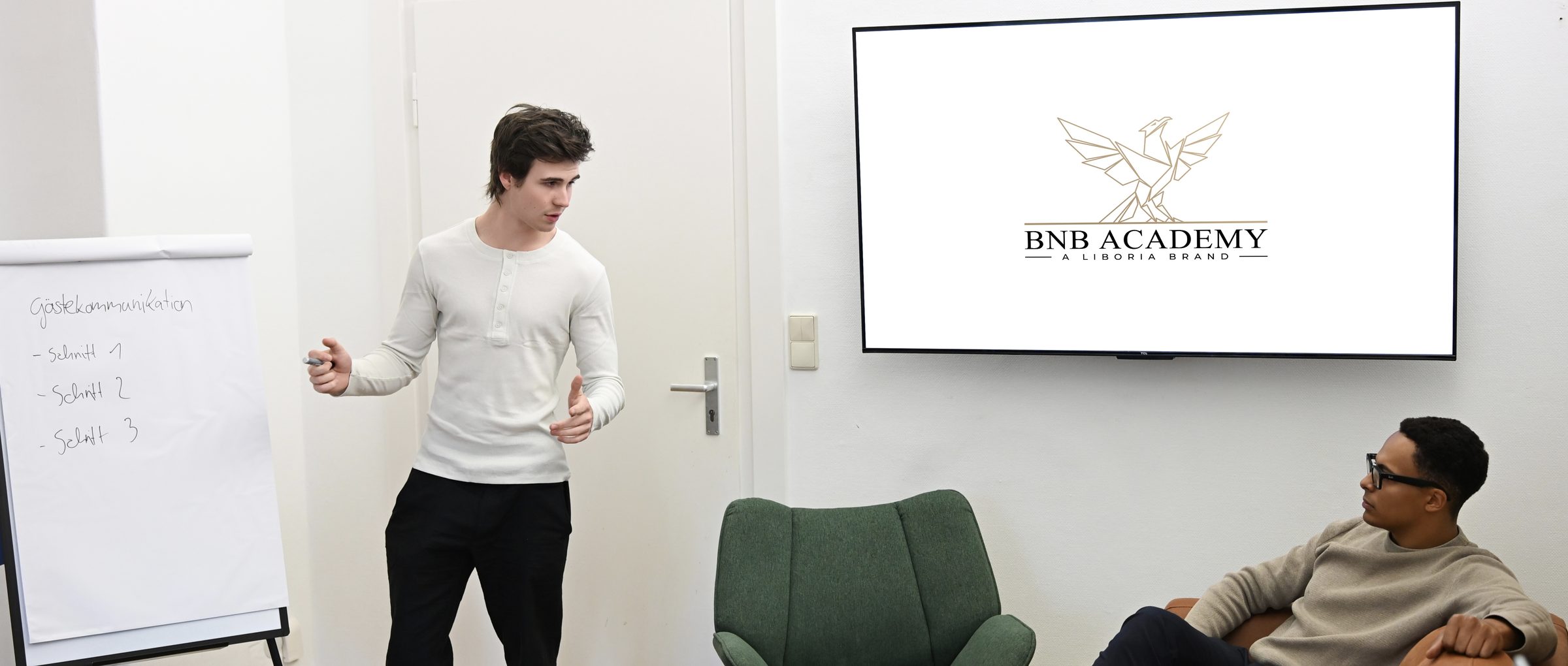 Laurin Henkel and Jomo Prill in the coaching room: one explains at the flip chart while the other sits in an armchair beside him; the BnB Academy logo on the screen behind them